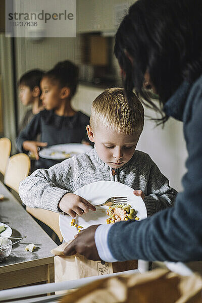 Männlicher Schüler wirft Essensreste in eine Papiertüte  die von einer Lehrerin in der Kindertagesstätte gehalten wird