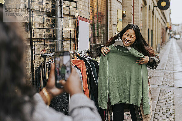 Reife Frau fotografiert fröhliche Freundin beim Anprobieren von Kleidung auf der Straße