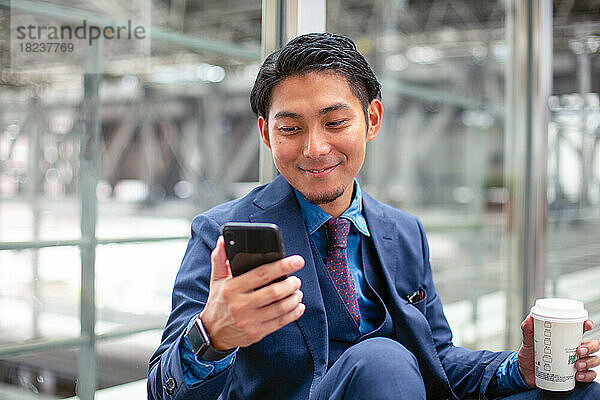 Ein junger Geschäftsmann im blauen Anzug in einer Stadt  der auf den Bildschirm seines Mobiltelefons schaut  eine SMS schreibt oder eine Nachricht liest.