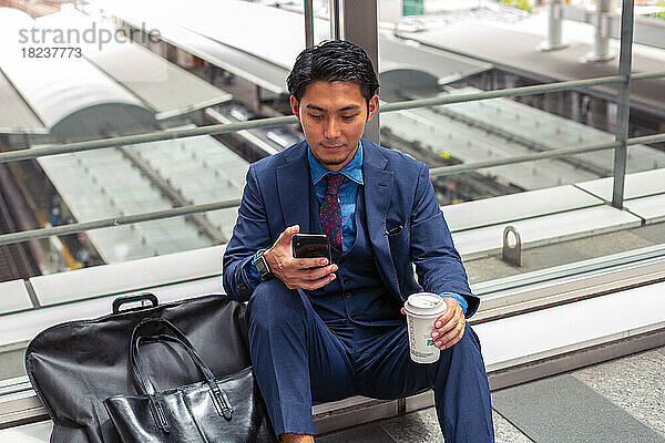 Ein junger Geschäftsmann im blauen Anzug in einer Stadt  der auf den Bildschirm seines Mobiltelefons schaut  eine SMS schreibt oder eine Nachricht liest.