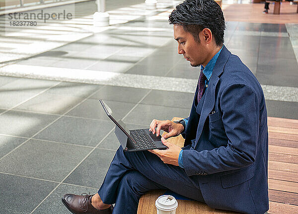Ein junger Geschäftsmann im blauen Anzug ist in der Innenstadt unterwegs und sitzt auf einer Bank und nutzt ein digitales Tablet.