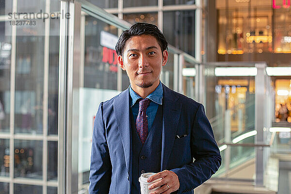 Ein junger Geschäftsmann im blauen Anzug ist mit einer Tasse Kaffee in der Innenstadt unterwegs.