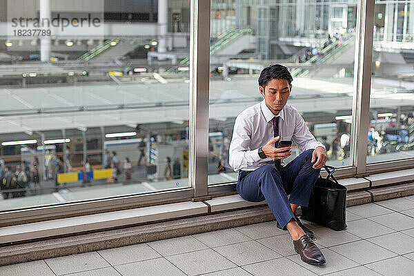 Ein junger Geschäftsmann in der Stadt  unterwegs  sitzt auf einem Gehweg und schaut auf sein Telefon.