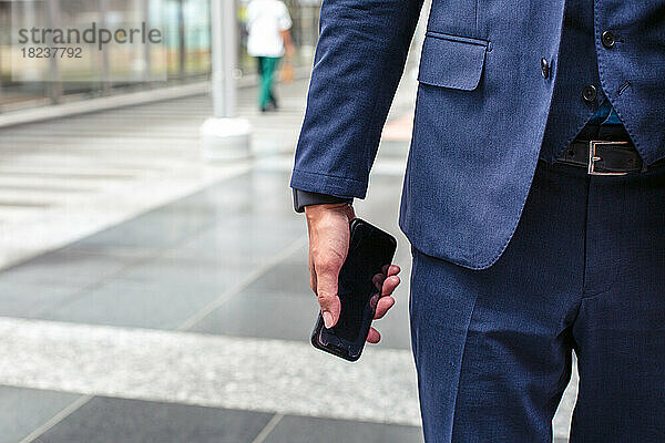 Ein junger Geschäftsmann im blauen Anzug ist in der Innenstadt unterwegs und hält sein Mobiltelefon in der Hand.