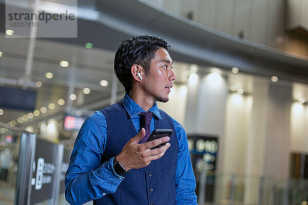 Ein junger Geschäftsmann in der Stadt  unterwegs  an einem Verkehrsknotenpunkt  hält sein Telefon in der Hand und schaut sich um.
