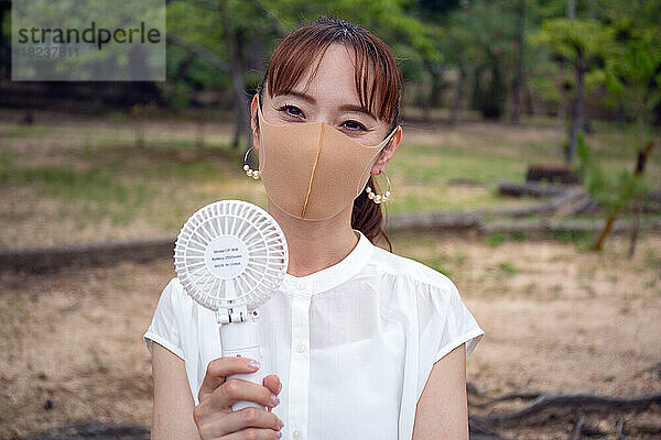 Eine reife Japanerin draußen in einem Park  trägt eine Gesichtsmaske und hält einen kleinen elektrischen Ventilator.