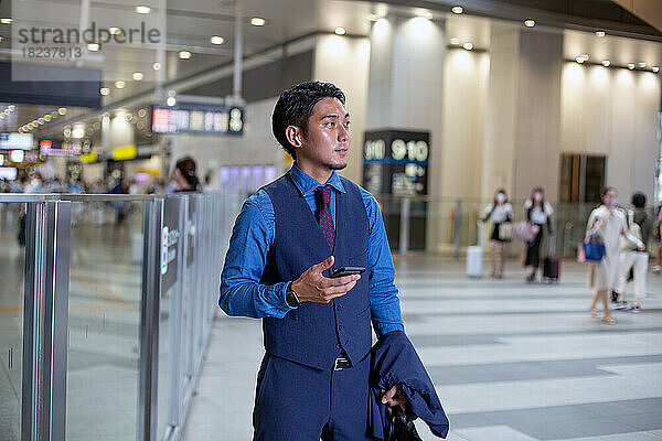 Ein junger Geschäftsmann in der Stadt  unterwegs  an einem Verkehrsknotenpunkt  hält sein Telefon in der Hand und schaut sich um.