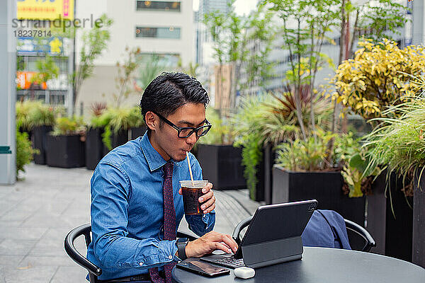 Ein junger Geschäftsmann in der Stadt  unterwegs  ein Mann  der draußen an einem Cafétisch sitzt  einen Laptop benutzt und mit einem Strohhalm an einem Erfrischungsgetränk nippt.