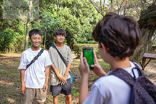 Ein Junge mit einem Mobiltelefon fotografiert zwei 13-jährige Jungen Seite an Seite  draußen in einem Park im Sommer.