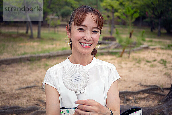Eine reife Japanerin steht an einem heißen Tag draußen in einem Park und hält einen kleinen elektrischen Ventilator in der Hand.