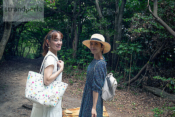 Zwei reife japanische Frauen in einem Park  die Taschen tragen  an einem Tagesausflug  die Köpfe gedreht  um in die Kamera zu lächeln.