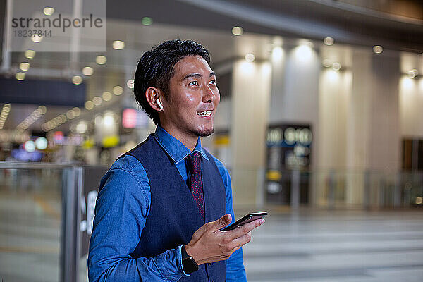Ein junger Geschäftsmann steht in der Stadt und schaut sich um  sein Mobiltelefon in der Hand.