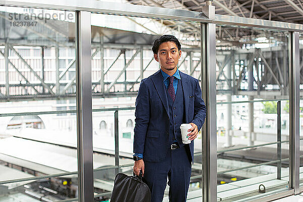 Ein junger Geschäftsmann im blauen Anzug ist mit einer Aktentasche und einer Tasse Kaffee in der Innenstadt unterwegs.