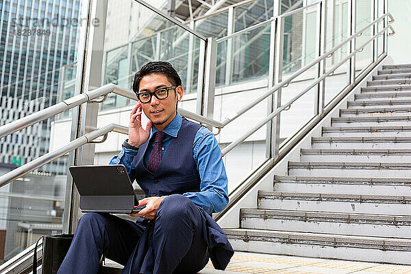Ein junger Geschäftsmann in der Stadt  unterwegs  sitzt auf der Treppe am Fenster und telefoniert  Laptop geöffnet.