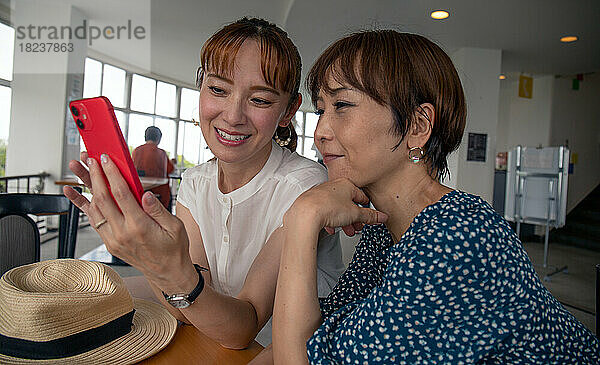 Zwei reife japanische Frauen  Freunde nebeneinander  sitzen und schauen auf den Bildschirm eines Mobiltelefons.