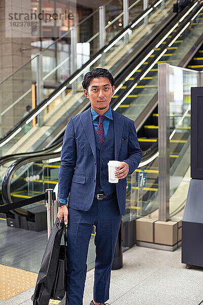 Ein junger Geschäftsmann im blauen Anzug ist mit einer Aktentasche und einer Tasse Kaffee in der Innenstadt unterwegs.