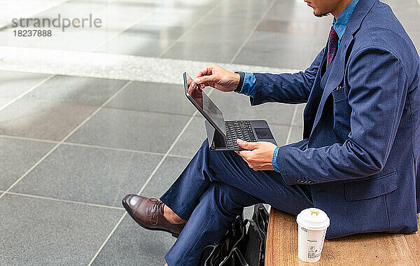 Ein junger Geschäftsmann im blauen Anzug ist in der Innenstadt unterwegs und sitzt auf einer Bank mit einem digitalen Touchscreen-Tablet.
