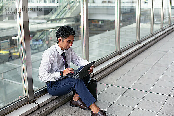 Ein junger Geschäftsmann in der Stadt  unterwegs  sitzt auf einem Gehweg und benutzt seinen Laptop.