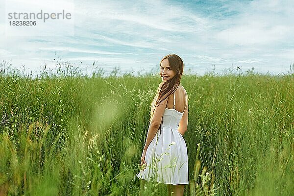 Fröhliche junge Frau in weißem kurzen Kleid auf dem Feld  die sich umdreht und über die Schulter schaut