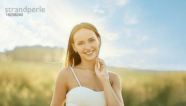 Porträt einer lächelnden jungen Frau im Spaghettiträgerkleid auf dem Feld mit der Sonne im Hintergrund