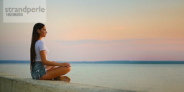 Ruhige junge Frau in Tanktop und Jeansshorts sitzt im Lotussitz auf dem Pier bei Sonnenuntergang