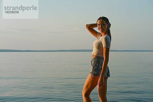 Glückliche junge Frau in weißem Tanktop und Jeansshorts am Strand bei Sonnenuntergang