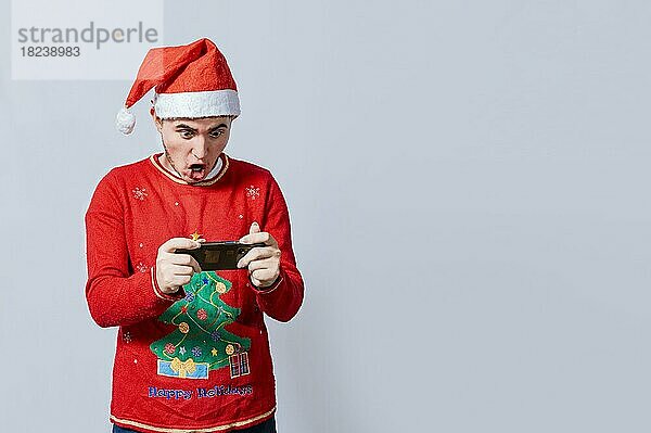 Mann mit Weihnachtsmütze sieht sich eine Weihnachtswerbung am Telefon an. Erstaunter Mann schaut sich eine Weihnachtswerbung am Telefon an  Erstaunter Mann mit Weihnachtsmütze schaut sich eine Weihnachtssonderaktion an