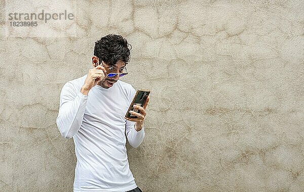 Überrascht junge Latin Blick auf sein Handy lehnt gegen eine Wand. Handsome Kerl mit Brille erstaunt mit seinem Handy lehnt gegen eine Wand  Latin Mann suchen Telefon lehnt gegen eine Wand