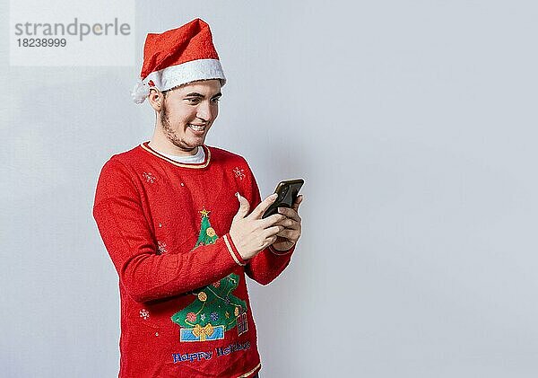 Menschen in Weihnachtsmütze mit und lächelnd auf Handy isoliert  Lächelnder junger Mann in Weihnachtsmütze mit Handy isoliert. Glücklicher Mann in Weihnachtsmütze lächelnd am Handy isoliert