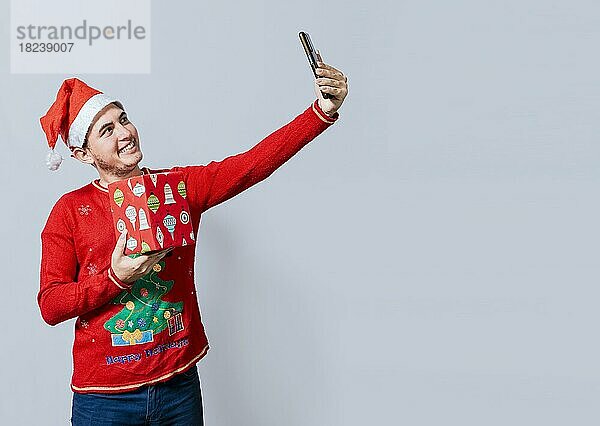 Weihnachten gut aussehend Mann  der ein Selfie hält ein Geschenk isoliert. Happy guy taking a christmas selfie isolated  Lächelnder junger Mann mit Weihnachtsmütze hält ein Geschenk und nimmt ein Selfie isoliert