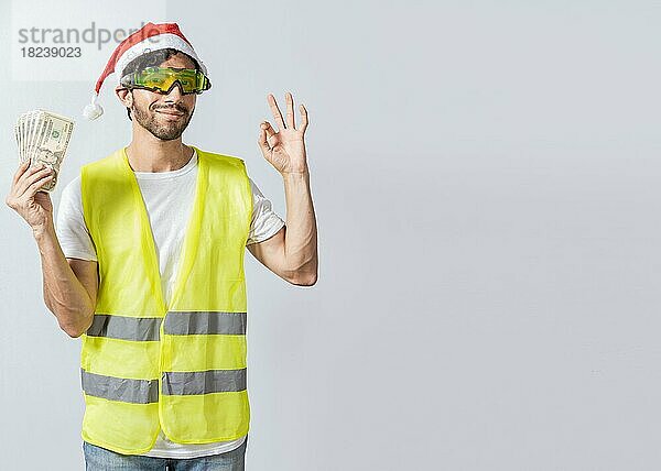 Ingenieur in Weihnachtsmütze hält Geld in ok Geste. Bauingenieur in Weihnachtsmütze mit zustimmenden Ausdruck hält Geldscheine. Weihnachten Ingenieur Konzept hält Geld in frohe Feiertage