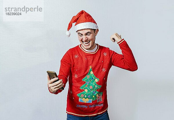 Teenager in Weihnachtsmütze feiert eine Promotion mit dem Handy  Glücklich lächelnder Mann in Weihnachtsmütze hält Handy feiern  Glücklicher Mann in Weihnachtsmütze hält Handy feiern