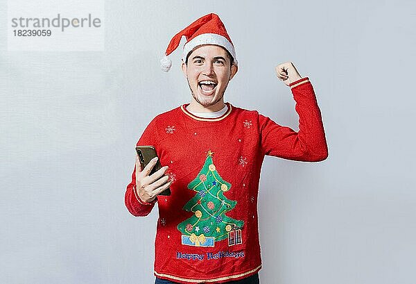 Glücklich lächelnder Mann mit Weihnachtsmütze  der ein Handy hält und feiert  Glücklicher Mann mit Weihnachtsmütze  Teenager mit Weihnachtsmütze  der eine Beförderung mit dem Handy feiert