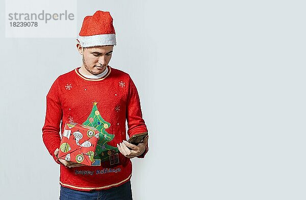 Mann mit Weihnachtsmütze hält Geschenkbox und lächelt auf Smartphone. Weihnachten Mann hält Geschenk-Box und Telefon isoliert. Handsome Mann in Weihnachtsmütze hält Geschenk und suchen Telefon