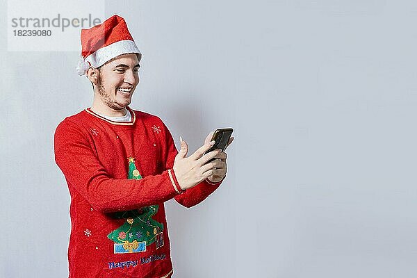 Glücklicher Mann in Weihnachtsmütze lächelnd am Handy isoliert. Menschen in weihnachtlichen Hut mit und lächelnd am Handy isoliert  Lächelnder junger Mann in weihnachtlichen Hut mit Handy isoliert