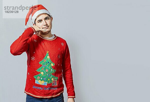 Mann in Weihnachtskleidung imitiert ein Telefongespräch  Junger lateinamerikanischer Mann in Weihnachtsmütze macht eine Anrufgeste isoliert. Menschen in Weihnachtskleidung machen einen Anruf Geste mit ihren Fingern