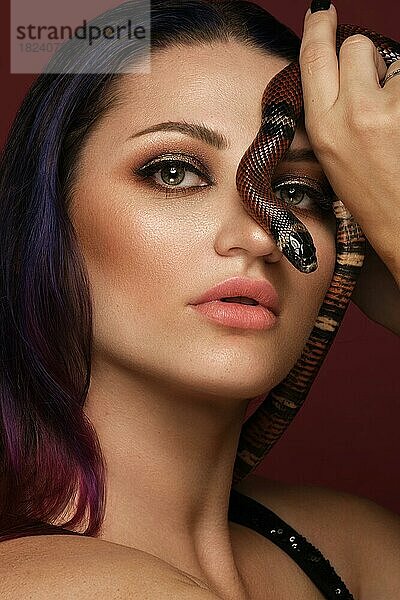 Schönes Mädchen brunette Modell mit Abendmode Make-up mit einer Schlange in ihren Händen. Schönheit Gesicht. Bild im Studio auf einem roten Hintergrund genommen