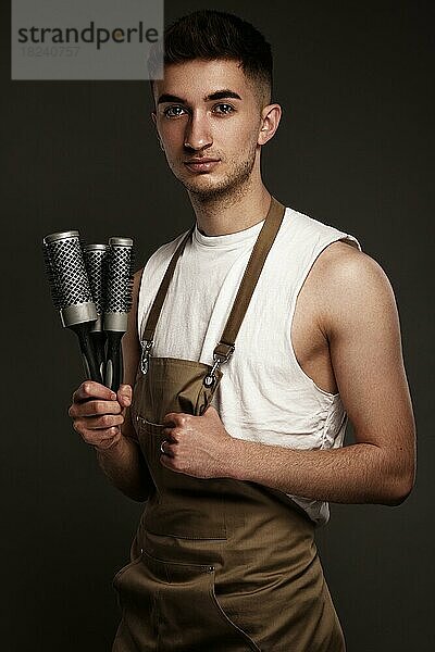 Gutaussehender Friseur in Arbeitsuniform mit Bürstenkämmen in den Händen. Foto im Studio aufgenommen