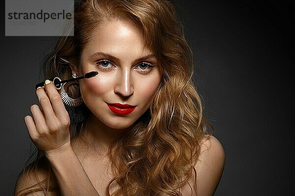 Schönes Mädchen mit roten Lippen und klassischen Make-up und Locken mit Wimperntusche in der Hand. Schönes Gesicht. Foto im Studio aufgenommen