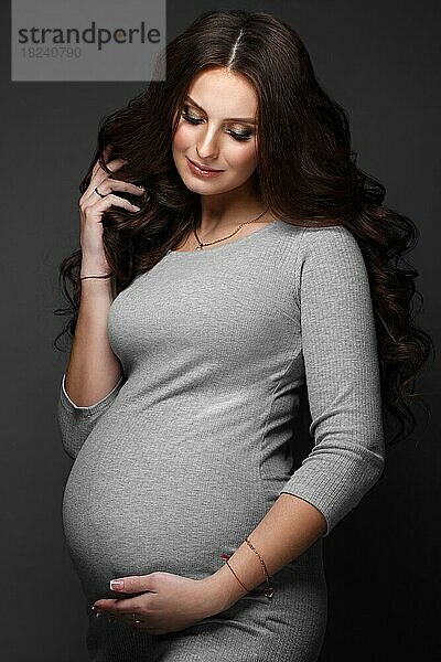 Schöne schwangere Frau in einem grauen Kleid  mit einem klassischen Make-up und Locken. Schönes Gesicht. Foto im Studio aufgenommen