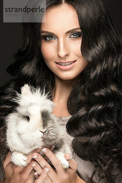 Schönes Mädchen brunette Modell  perfekte Locken und Abendmode Make-up mit einem Kaninchen in ihren Händen. Schönheit Gesicht. Bild im Studio auf einem schwarzen Hintergrund genommen