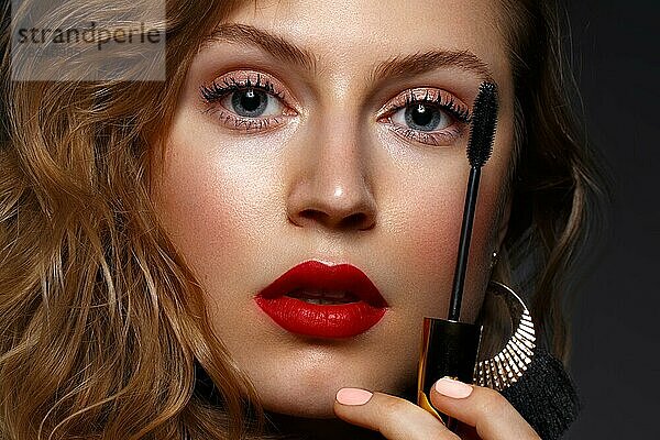 Schönes Mädchen mit roten Lippen und klassischen Make-up und Locken mit Wimperntusche in der Hand. Schönes Gesicht. Foto im Studio aufgenommen