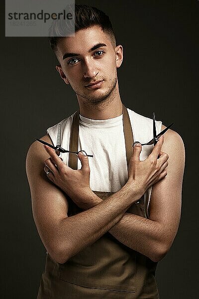 gutaussehender Friseur in Arbeitsuniform mit einer Schere in der Hand. Foto im Studio aufgenommen