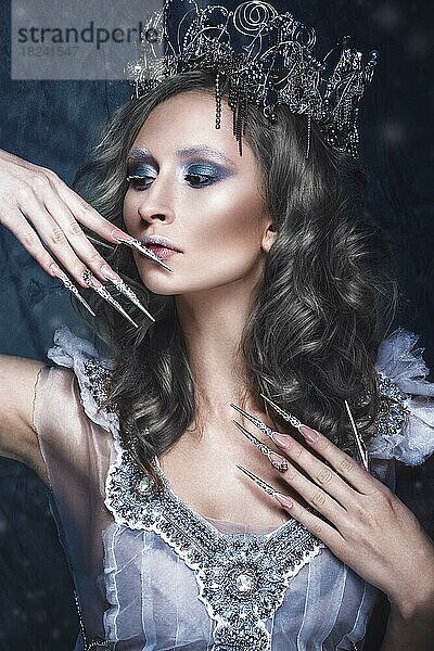 Schönes Mädchen im Bild der Schneekönigin mit kreativem Make-up in einem transparenten Kleid mit einer Krone und einer langen Nagelkunst. Die Schönheit des Gesichts. Fotos im Studio geschossen