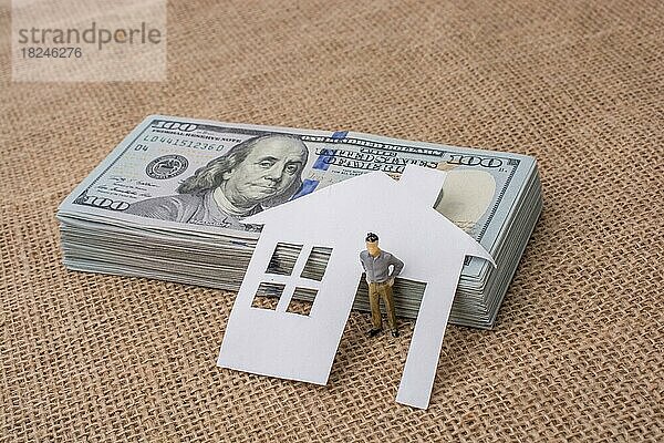 Haus aus Papier und eine Männerfigur neben einem Bündel von US-Dollar-Banknoten