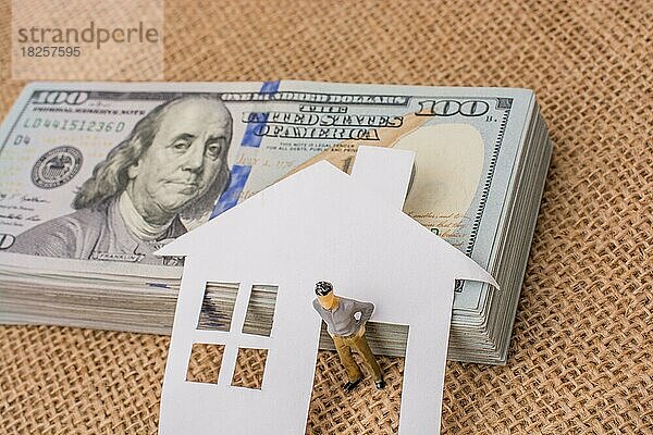 Haus aus Papier und eine Männerfigur neben einem Bündel von US-Dollar-Banknoten
