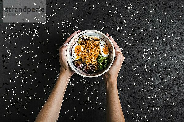 Erhöhte Ansicht Hände halten Schalen Ramen-Nudeln mit Eiern Salat Ausbreitung mit Reiskörnern schwarzen Hintergrund