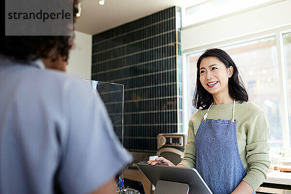 Lächelnder Cafébesitzer mit Schürze im Gespräch mit dem Kunden an der Kasse