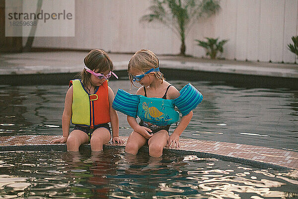 Zwei Kinder in Schwimmanzügen und Schutzbrillen sitzen zusammen am Pool