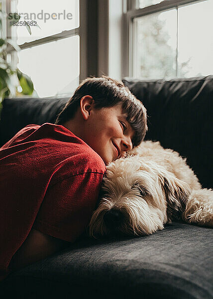 Glücklich lächelnder Junge kuschelt mit flauschigem Hund auf einem Sofa zu Hause.
